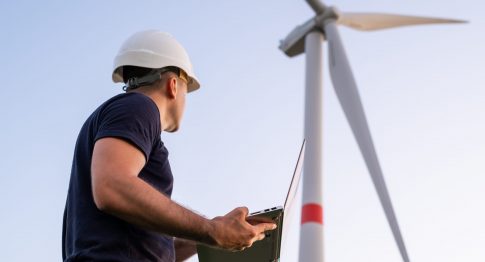 Male engineer in the helmet controls and maintains the operation of a wind turbine using a laptop on the background of windmill.